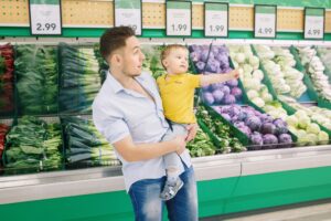 Man holding a baby in a grocery store with price tags above produce